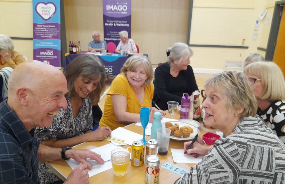 Quiz team sat at a table smiling with quiz hosts in the background. Imago banners on display.